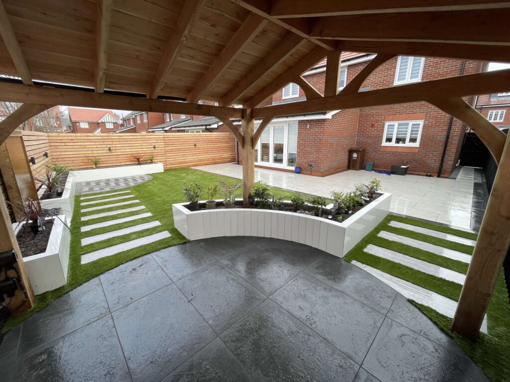 Oak pergola over dark porcelain with curved white raised planters and artificial grass