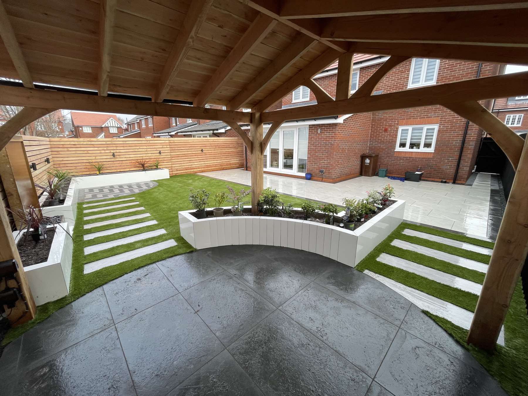Oak pergola over dark porcelain with curved white raised planters and artificial grass